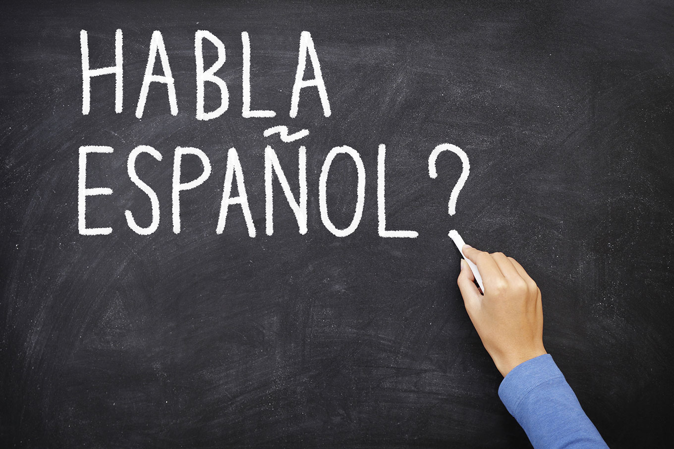 Imagem de um quadro negro com uma mão segurando um giz e escrevendo "Habla español?", em referência ao curso de espanhol